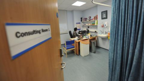 A GP sits at his desk in a consulting room at a surgery. A sign on the door, which is partially open to allow a view into the room, says "consulting room 1".