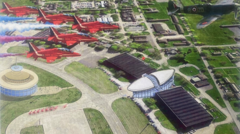 An artist's impression showing a bird's eye view of a former RAF base, with new buildings and older structures standing side by side. At the top of the image, a squadron of red jets of the Red Arrows are flying overhead behind a World War Two Spitfire fighter plane. On the ground, the old roads of the base can be seen, with grass in between. To the left, people are walking up to a round building, made largely of glass. In the centre another glass building has two large, black warehouse-like wings.