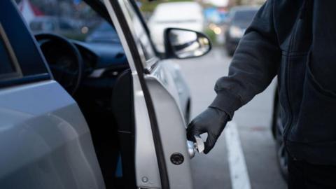 Shows a man with a black glove and black coat opening a silver-coloured car