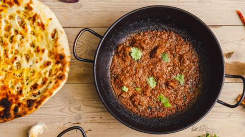 a balti curry in a black pan next to a naan bread on a wooden table