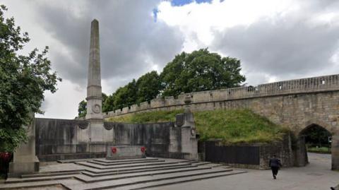 The North Eastern Railway War Memorial - a tall, grey-stone obelisk surrounded by a three-sided screen wall, which includes the names of thousands of war dead. It stands next to the York city wall - an ancient stone structure with battlements and an archway. 