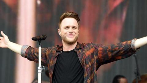 Olly Murs standing on stage, with his arms out stretched. There is a microphone in front of him, with a white stand. Murs has brown short hair and is wearing a orange, black and pink patterned shirt over a white t-shirt. He is smiling. 