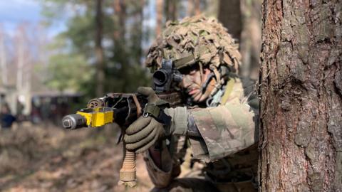 UK soldier on Nato mission in Poland - he is in camouflage clothing, holding a gun and standing next to a tree