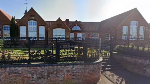 The exterior of Fakenham Junior School, behind a brick wall topped with a decorative metal fence. It is a sunny day.