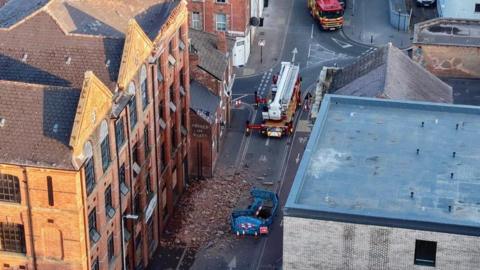 Aerial view of an urban street with an old brick warehouse building, rubble in street and fire engines in attendance