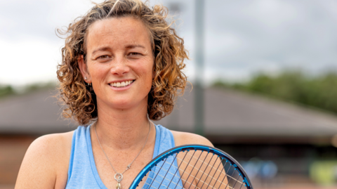 A professional close-up shot of Catherine, smiling to camera, wearing a blue sports vest and holding her tennis racket up near her shoulder.  She has very curly brown hair. The background of the tennis court is blurred.