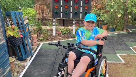 11-year-old Jenson is sitting in his power chair smiling at the camera. He is sat in his new garden which has a new path and F1-style racing lights.