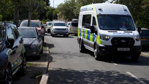 A convoy of police vehicles travelling through a residential street on a sunny day.