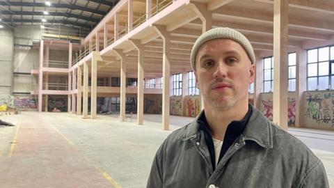 A man in a white woolly hat and black jacket stands in a huge warehouse.