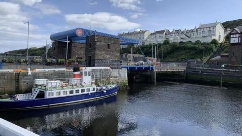 The lifting bridge over Douglas Harbour. It is stone built with a blue lifting mechanism. A long passenger boat is moored against the harbour wall in front of it.