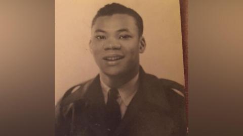 A black and white photo of a young man with short brown hair smiling for the camera and wearing a dark blazer, tie and light shirt. He looks very smart and is in uniform.