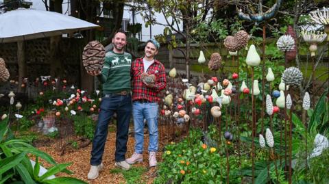 A man in a green Christmas jumper and dark trousers is standing next to a man in a checked red flannel shirt and jeans. They are standing in a garden.