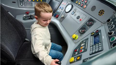 Five-year-old Daniel Rogerson from Wigan sits in the driver's cab of one of his favourite trains. He is operating some of the buttons while sitting in the driver's seat. Daniel wears a cream jumper and blue denim jeans. 