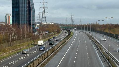 Three lines of road. The central lane has a few vehicles in the distance and the other two are busier. There are a number of electric pylons and some buildings on the left-hand side of the road. 