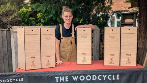 Yana is standing behind a table with five presentation boxes lined up on it. She has light brown hair tied up in a bun and is wearing glasses and is smiling a the camera. She is wearing and a black t-shirt and beige apron and is resting her left arm on one of the boxes.