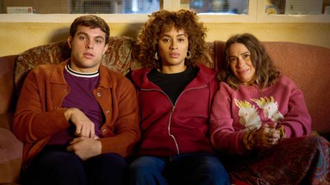 Three people sat on a couch looking at the camera