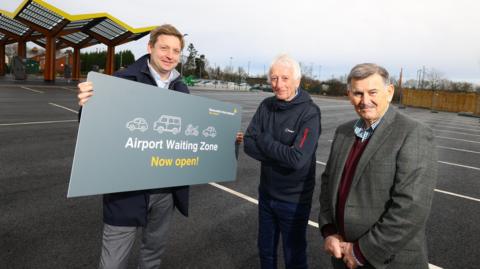 Dean Ward, Director of Commercial at Newcastle Airport, John Littleton, of Woolsington Parish Council, and Mayor of Ponteland, Alan Hall at the new waiting zone. Dean Ward is holding a grey sign with various vehicles painted on it reading Airport Waiting Zone Now Open! He has blue eyes and blonde hair and is wearing a blue shirt and navy blue coat with grey trousers. John Littleton has short white hair. His arms are crossed. He is wearing jeans and a black jacket. Alan Hall has short grey hair and a moustache. He is wearing a grey tweed suit jacket, a red vest and blue square pattern shirt.