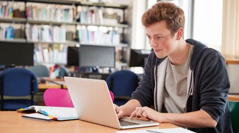 Male GCSE student working at laptop in school library revising with past papers