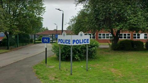 A picture of a police station. There is a white sign in the grounds of the station, that has blue letters spelling police. Behind is a large green tree and behind that is the station.