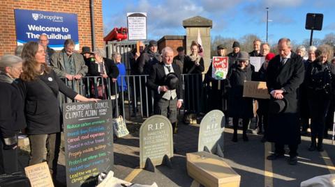 A gathering of people stand outside one corner of Shropshire Council's headquarters in Shrewsbury. They are dressed in black and some are holding placards, while others are stood behind mock coffins and headstones that are on the floor.