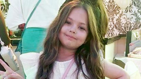 Olivia Pratt-Korbel, with long brown hair and a white T-shirt on, smiles and holds her hand in a peace sign as she looks at the camera in a busy room