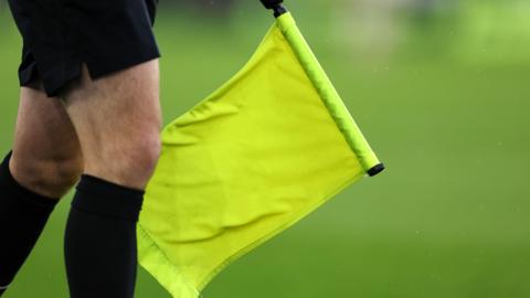 The legs of a football referee who is wearing black socks and holding a yellow flag. The grass is blurred in the background. 