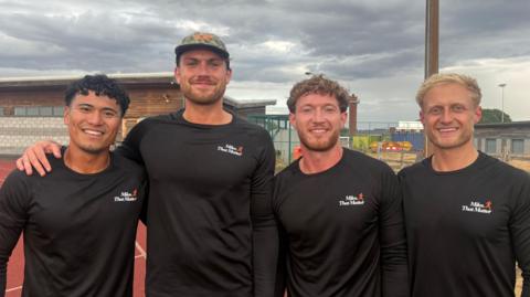 Four men standing on a running track. They are all wearing long-sleeve black tops with a white and orange Miles That Matter logo on the chest. The man on the left has black curly hair. The man next to him has brown facial hair and is wearing a camouflage cap. Next to him is a man with brown curly hair and facial hair. The man on the right has short, blond hair. The sky is cloudy and grey.