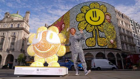 Mr Doodle stands beside one of his sculptures in Piccadilly Circus. The installation features his signature black-and-white doodle artwork with bright orange and yellow colours, while an image of the artist appears on the famous Piccadilly Circus screen behind him