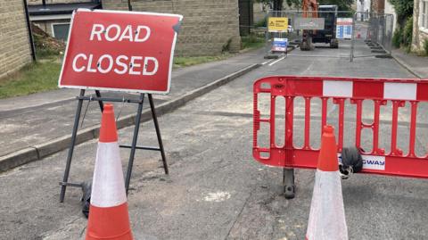 A photo showing a road with a red road closed sign, two red and white comes and a red barrier. In the background is a fenced off area containing a digger