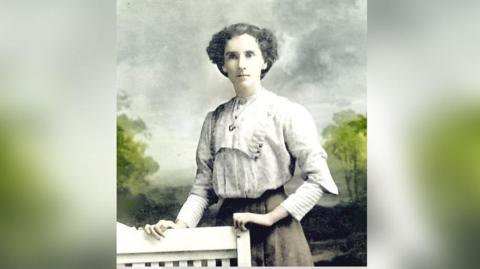 A lady stands holding onto a chair in what appears to be a very old photograph. She is wearing a blouse and skirt and is in a photography studio with a tree lined background behind her