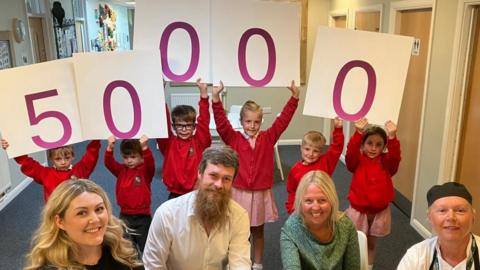 Burton Green Primary School children hold up individual signs with numbers to represent 50,000 school meals have been served as part of the Hungry Minds Appeal scheme. Staff members and councillor Bob Webb sit in front of the children. 