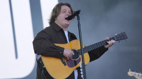Lewis Capaldi performing on the Pyramid Stage during the Glastonbury Festival at Worthy Farm in Somerset in 2025. He stands in front of a microphone as he plays the guitar.