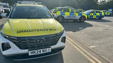 Emergency service vehicles are parked in a group, three police cars and one ambulance service car and one car marked HM Coastguard.