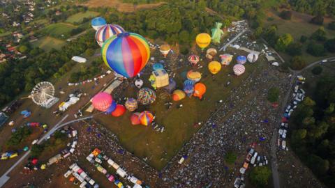 Dozens of hot air balloons are seen taking off from the Bristol International Balloon Fiesta at Ashton Court. They are many different colours and some are in the air already but many are inflated but still on the ground. The trees and grassy areas of Ashton Court are also visible in the photo, which is taken from a balloon which has already taken off, along with hundreds of spectators. There is also a ferris wheel-style ride visible