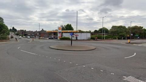 The small roundabout with a Shell petrol station at the back. A number of cars in the distance are travelling towards the junction. 