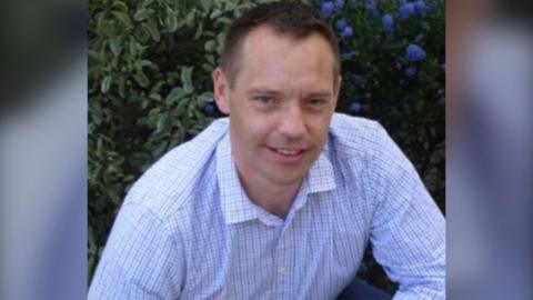 Scott Wilson smiling at the camera. He is wearing a blue checked shirt and is crouching in front of a green bush that has blue flowers on the right of it.