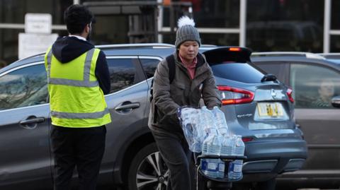A woman is pushing a trolley full of water bottles through a car park. 
