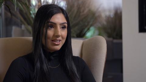 British Asian woman sitting on a cream sofa. She has black long hair and wearing a black turtle neck jumper with a gold thin chain around her neck. There are plants in the background.