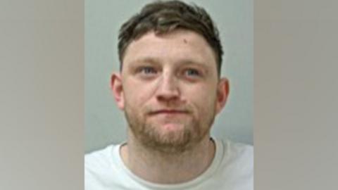 Police photo of Daniel Burba, who has short brown hair and a brown stubble-beard and is wearing a white T-shirt