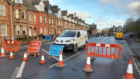 A residential road is shut off by a police cordon and orange blockages. Several vans are parked within the cordon. Houses line the left hand side of the image. 