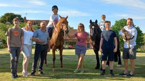 Two families of four, which includes two children on horseback, are stood on a lawn in front of trees and a blue sky
