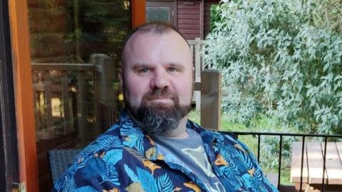 Mark Willerton in his wheelchair on a balcony, wearing a summery short-sleeved blue shirt and grey shorts.