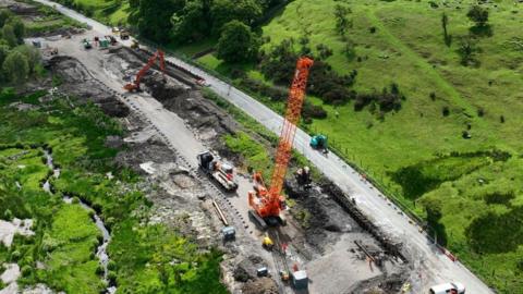 Two orange cranes work to repair a road with green fields on either side