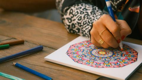 A person draws a colourful round pattern using pencils and pens. They are wearing a white and black animal print shirt and gold rings.