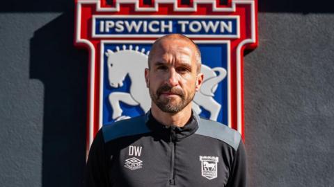 David Wright is looking at the camera, standing in front of the Ipswich Town football crest. He is balding with a beard and moustache. He is wearing a dark top which also has the club logo on it.