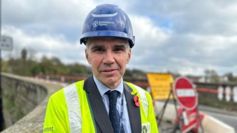 A man in a yellow hi-vis jacket and blue helmet with the words Environment Agency printed in white across it. He is wearing a grey blazer jacket, blue shirt and dark blue tie. He has a red poppy on his right lapel.