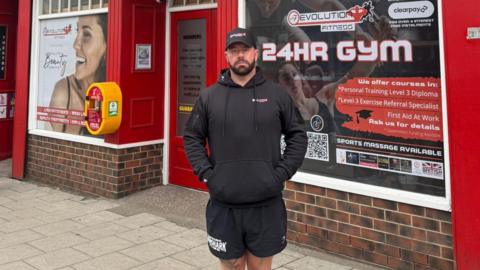 Micky Donkin is standing in front to the Evolution Fitness Gym which has a red painted exterior with various stickers in the window, including one that says it is a 24-hour gym. Mr Donkin has a black beard and is wearing a black cap, hoody and shorts.