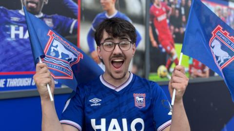 Jack Richardson is holding two flags either side of him. He has a happy expression and has short brown hair. He is wearing black framed glasses and a blue Ipswich Town FC football T-shirt.