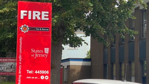 A large red sign outside of a fire station. The sign says Jersey Fire and Rescue. States of Jersey. It is located in front of a tree and a large building.