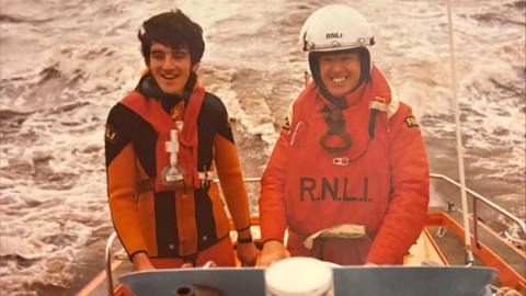 An old and slightly sepia toned photograph of Richard in a red RNLI uniform on a speed boat with a colleague. He is on the left and smiling. His colleague who is also wearing uniform, is wearing a white helmet and smiling.